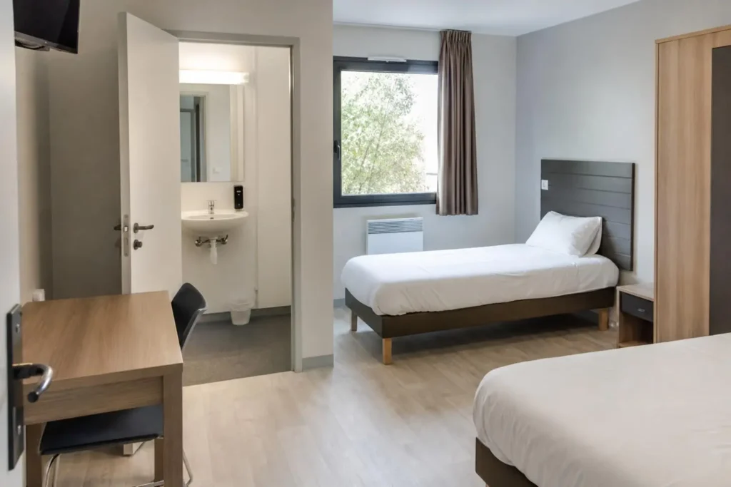 Chambre d’hôtel propre et lumineuse avec deux lits simples, bureau et chaise, penderie, baie vitrée et salle de bains attenante. Intégration naturelle: hébergement workers détachés france.